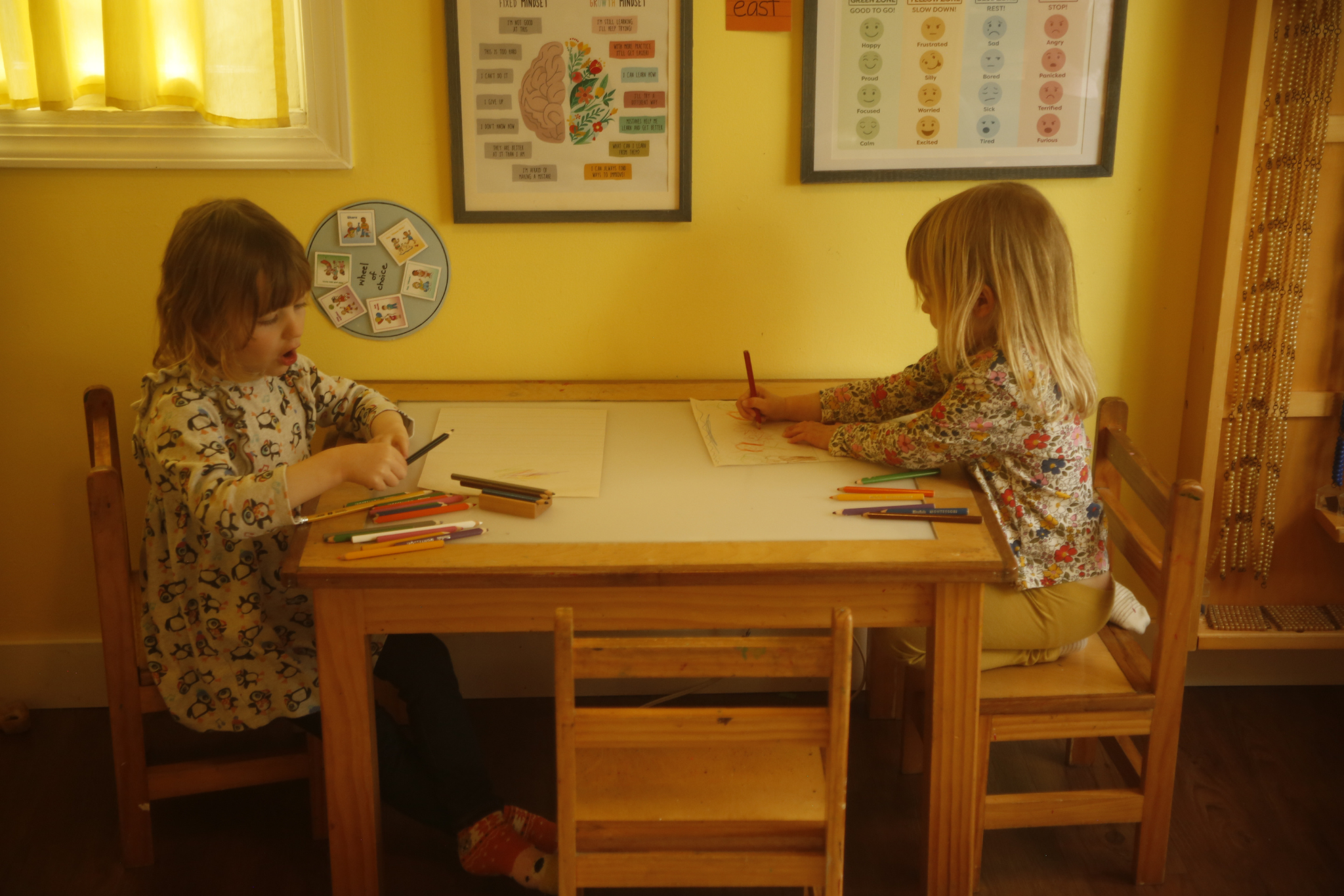 Two children drawing with colored pencils at a small wooden classroom table