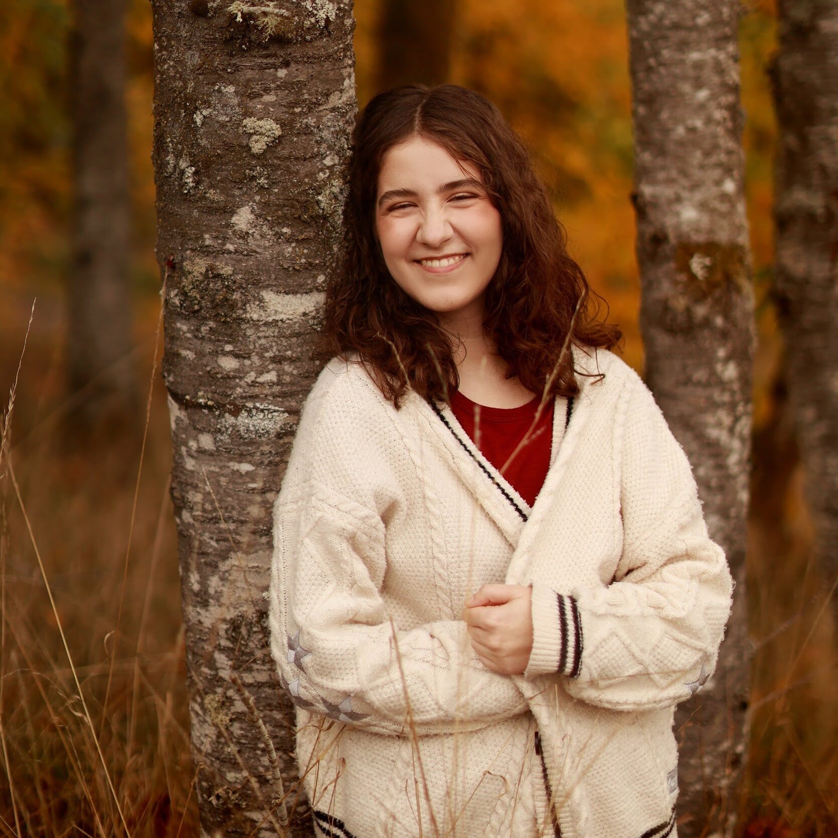 Smiling young woman in a cream sweater standing among trees with autumn leaves
