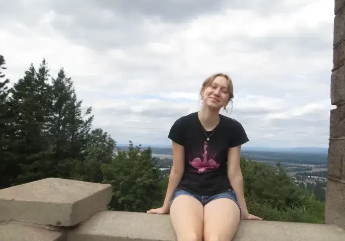 Smiling young woman sitting on a stone wall with trees and a scenic view behind her